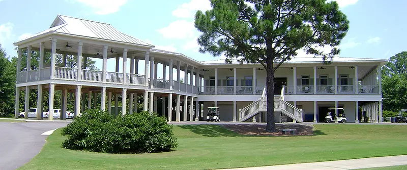 The clubhouse verandas and white columns behind a mown lawn, with carts parked underneath and a tall pine alongside