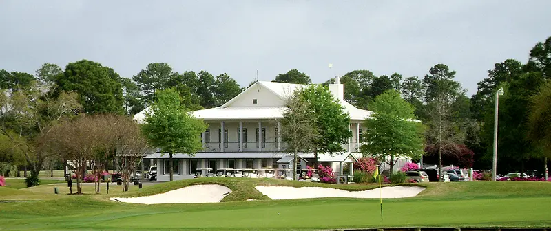 The clubhouse seen from across a green, with a yellow flagstick, bunkers and flowering azaleas in front of it