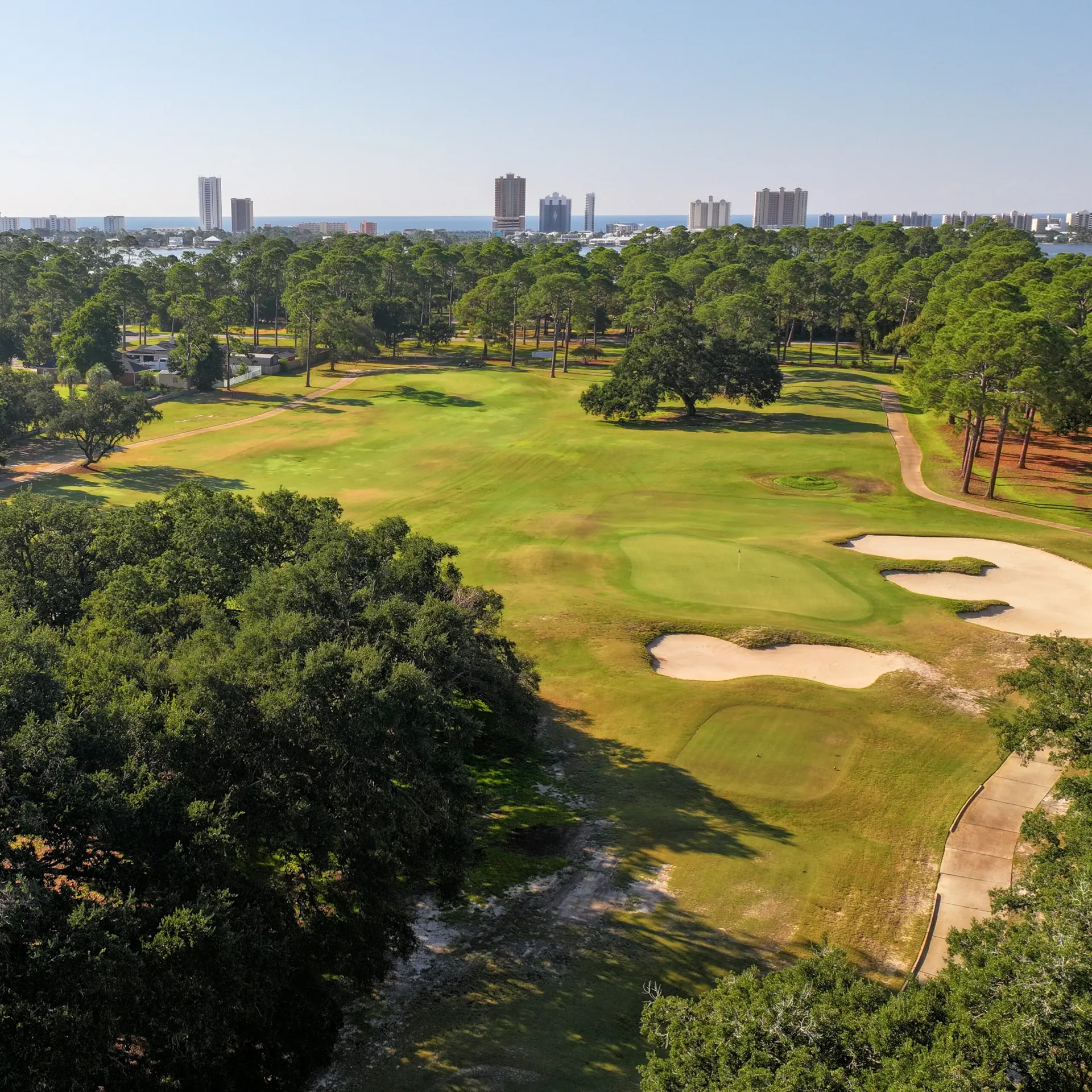 Aerial view along a tree-lined fairway to a bunkered green, with the Gulf Shores skyline and the Gulf beyond