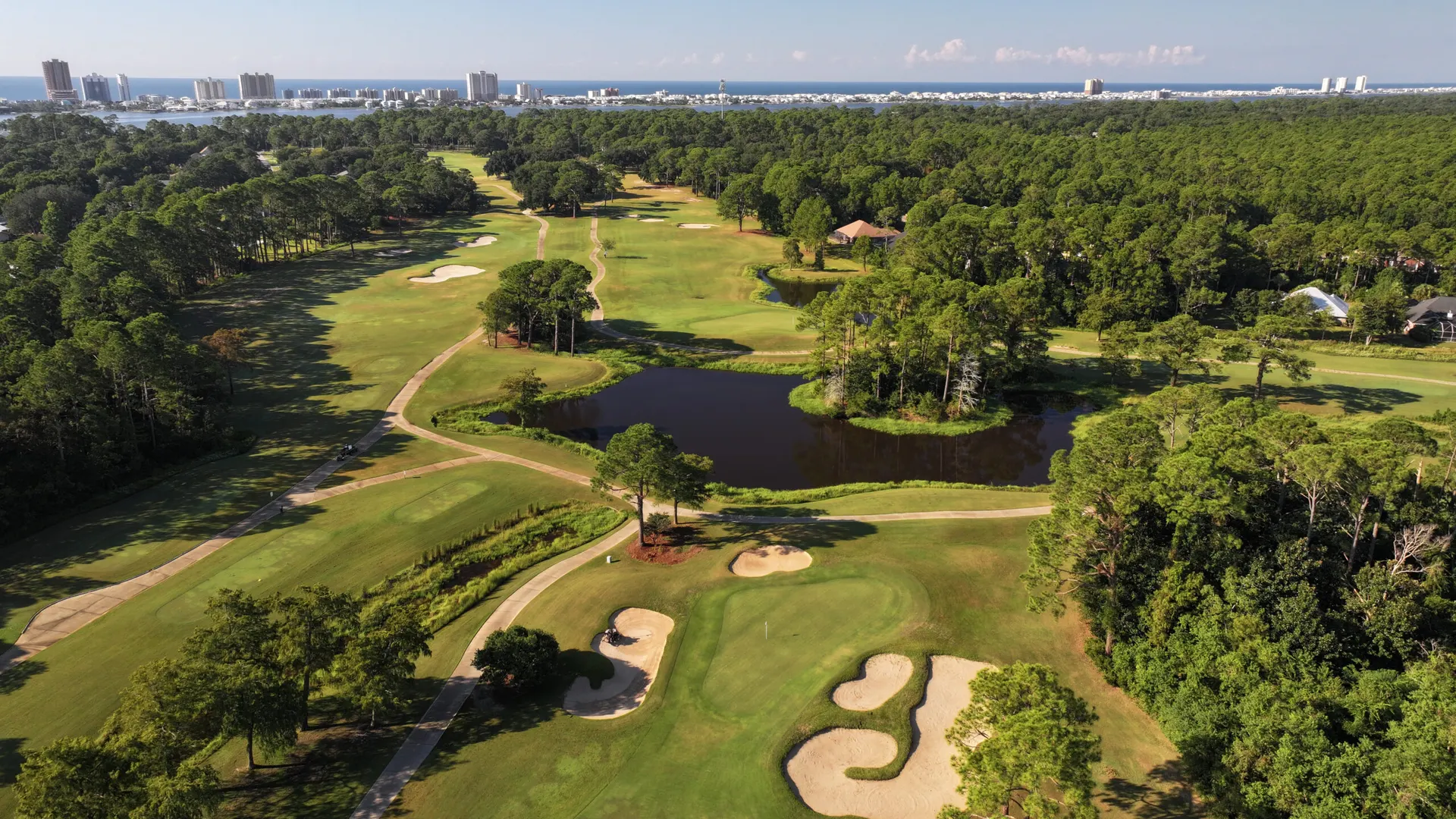 Aerial view of the course with a lake, bunkered greens and cart paths winding through pines, the Gulf on the horizon