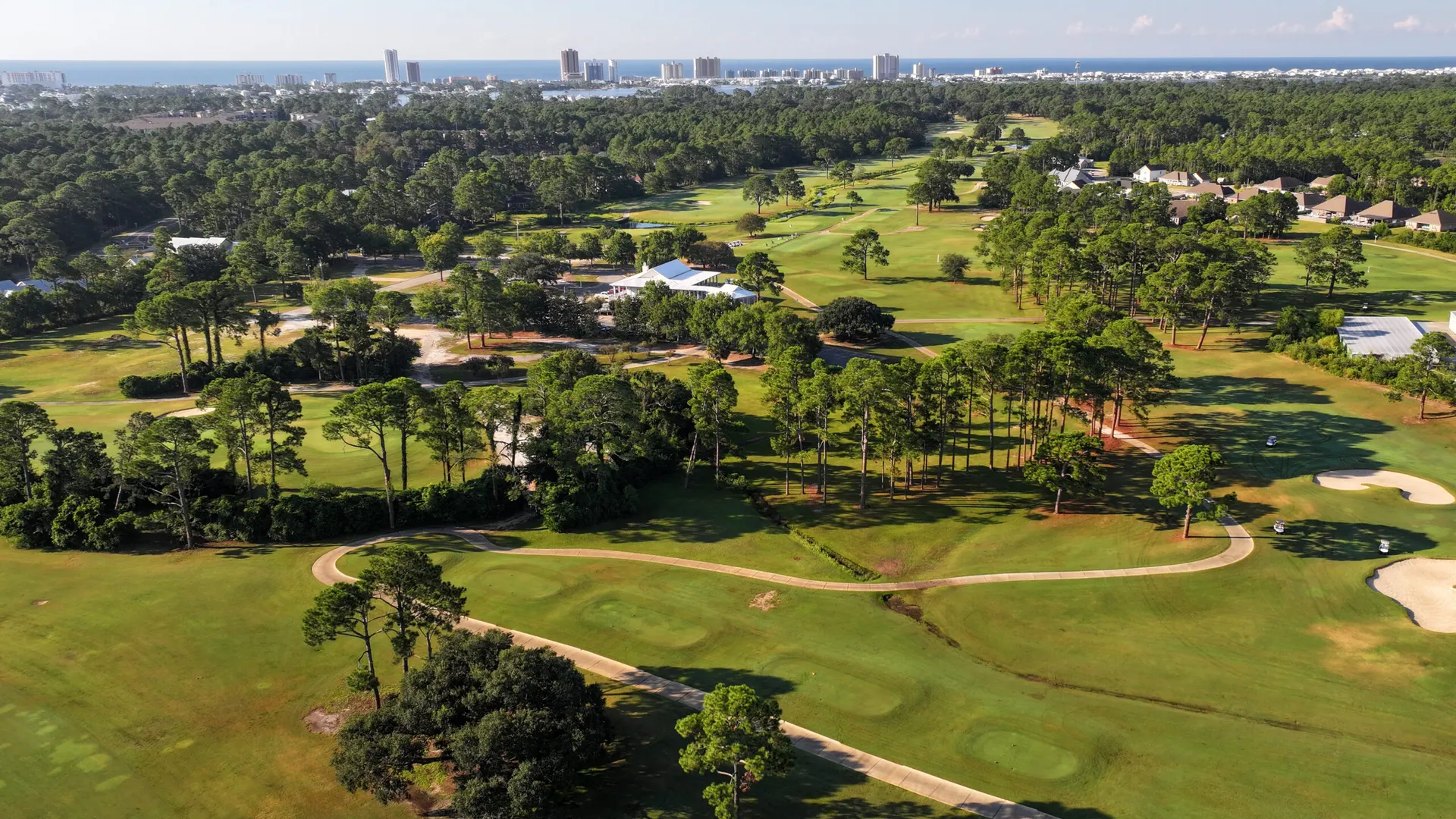 Aerial view of fairways and pines around the clubhouse, with the Gulf Shores skyline and the Gulf on the horizon