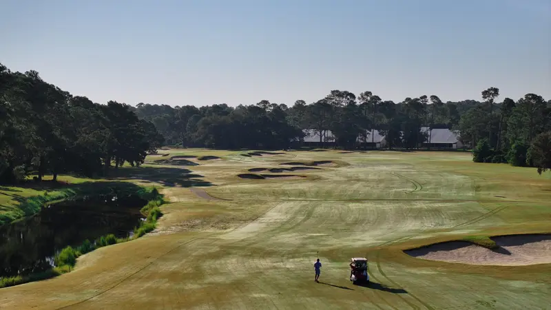 A golfer walking beside a cart down a wide fairway in early morning light