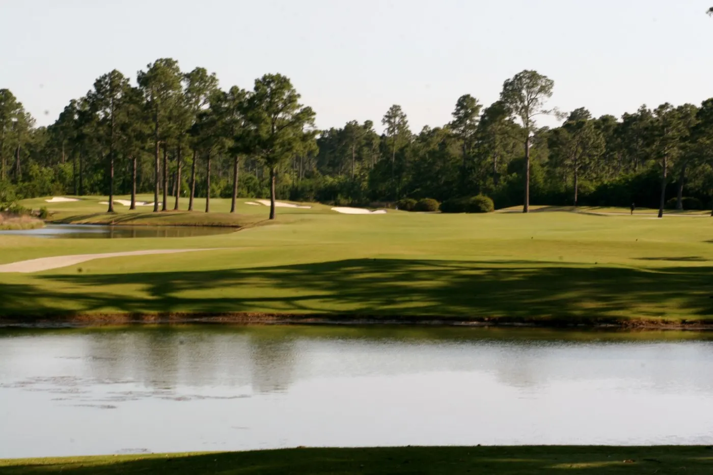 Lake in the foreground with a bunkered fairway and pine woods beyond at Gulf Shores Golf Club