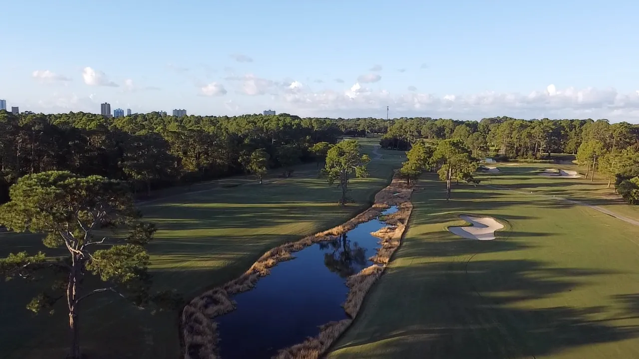Aerial view down a fairway split by a reed-lined creek at Gulf Shores Golf Club, with the Gulf Shores skyline beyond the pines